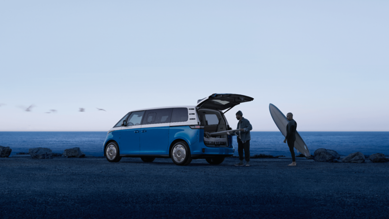 The VW ID. Buzz 2025 is parked by the beach, with two people loading a surfboard into the back. 