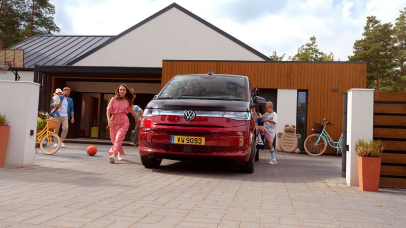 Van Volkswagen Multivan rouge stationné devant une maison, avec des personnes sortant du véhicule et des objets du quotidien autour.