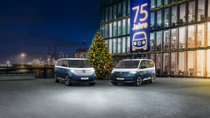 Ein blauer und weißer Volkswagen ID. Buzz und ein blauer und weißer Volkswagen Multivan stehen nachts vor einem festlich beleuchteten Gebäude mit einem großen "75 Jahre" Banner und einem Weihnachtsbaum.