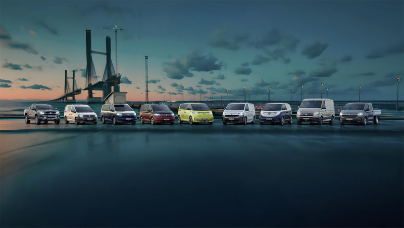 Cinco vehículos comerciales Volkswagen de diferentes modelos y colores, incluyendo una furgoneta camper y una furgoneta eléctrica ID.Buzz, alineados al aire libre al anochecer con un edificio moderno y un puerto de fondo. La imagen representa la diversidad de la gama de vehículos comerciales de Volkswagen.