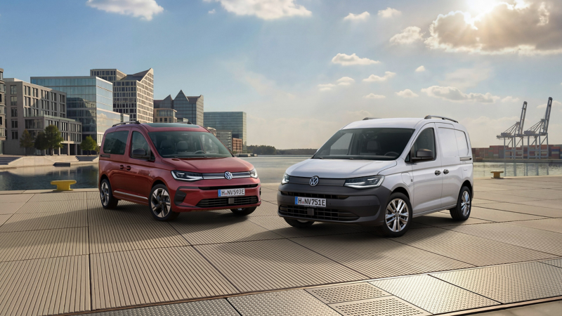 Ein roter VW Caddy Style und ein weißer VW Caddy Cargo stehen nebeneinander auf einem gepflasterten Hafengelände vor einer modernen Stadtkulisse.