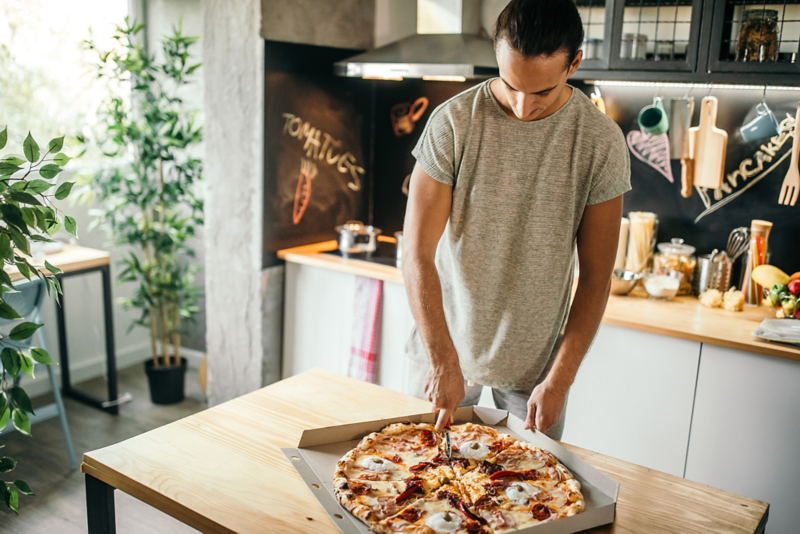 En mand skærer en pizza ud i køkkenet.