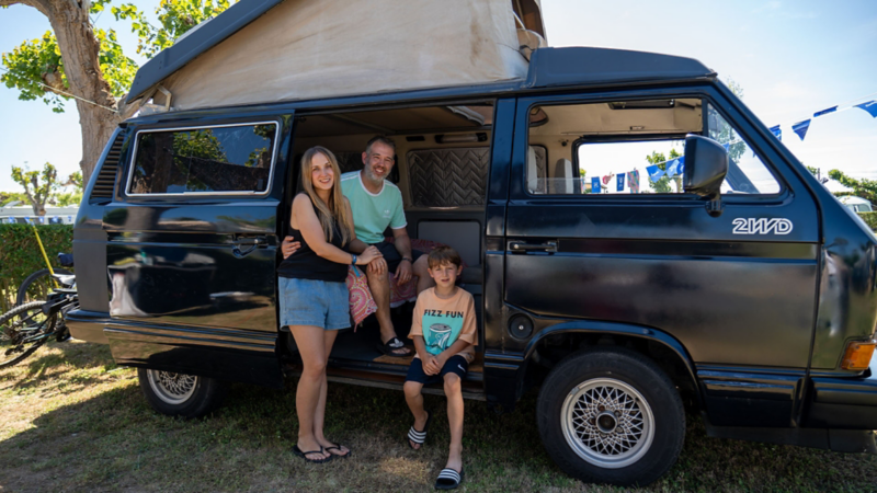 Una familia sentada en el lateral de un VW T3 con las puertas abiertas.