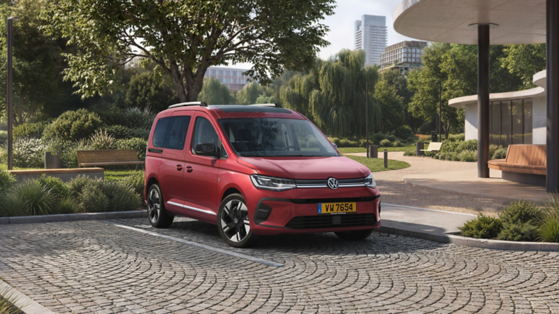 Volkswagen Nouveau Caddy rouge stationné dans un espace urbain verdoyant, sur une place pavée entourée d’arbres et de bâtiments modernes.