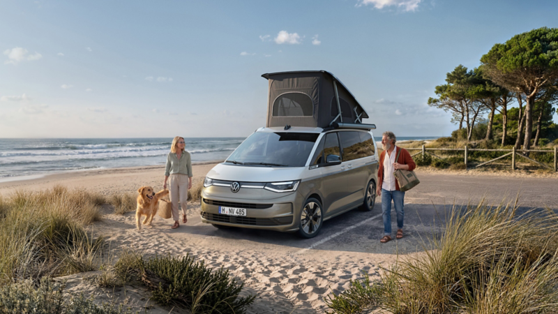 Volkswagen California avec toit relevable ouvert stationné près d’une plage. Deux personnes et un chien se promènent à côté du véhicule, avec l’océan et les dunes en arrière-plan.
