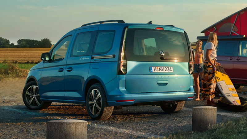 Un van Volkswagen Caddy california bleu est garé sur un chemin de terre. Deux femmes, dont l'une tient une planche de surf, se tiennent à l'arrière du véhicule.