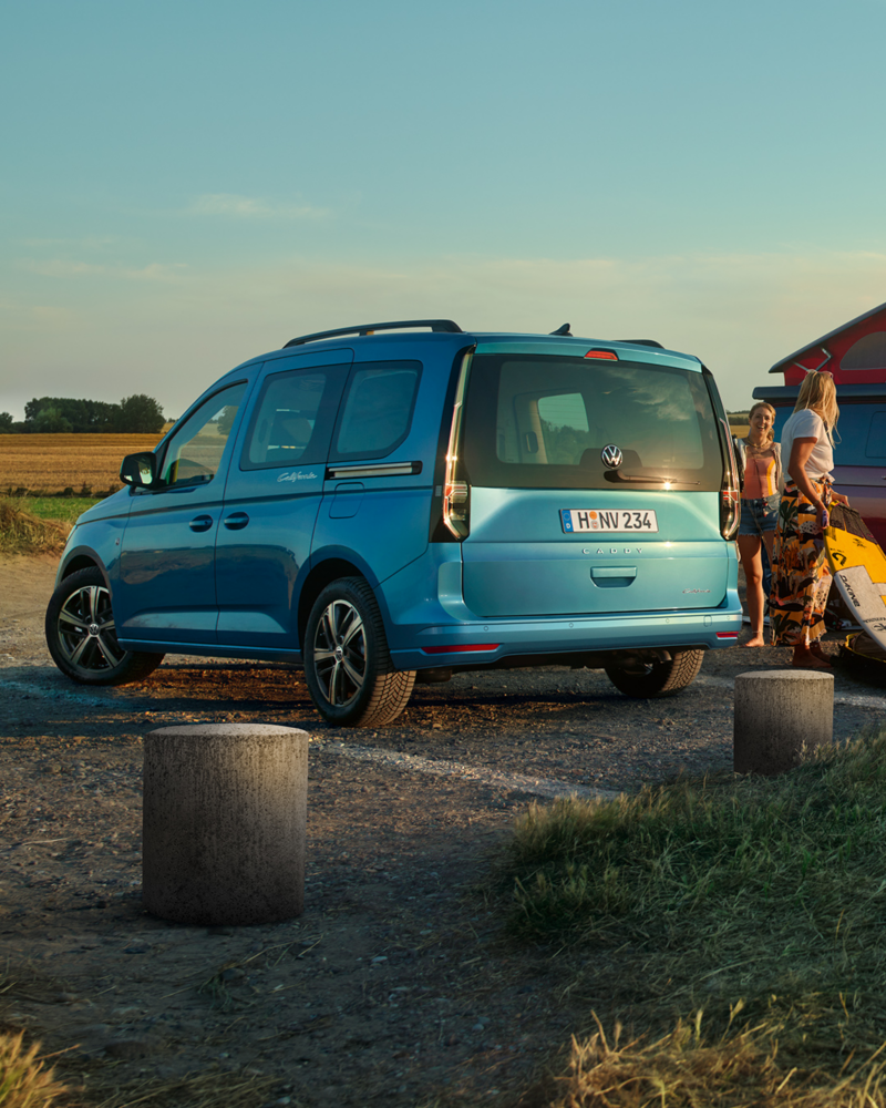 Un van Volkswagen Caddy california bleu est garé sur un chemin de terre. Une femme se tient à l'arrière du véhicule, devant un paysage de champs.