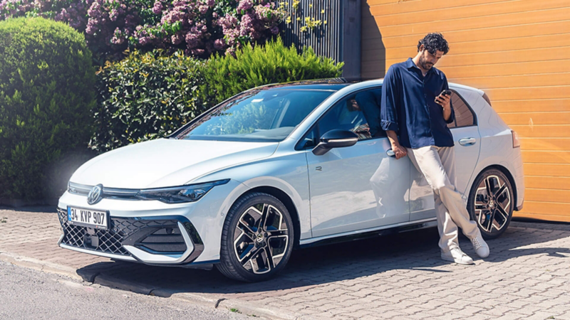 A person using a phone and leaning against a white Golf in a driveway