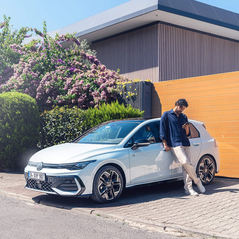 A person using a phone and leaning against a white Golf in a driveway