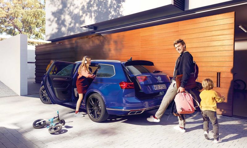 Kind steigt in VW Passat Variant ein, Mann trägt Taschen und nutzt seinen Fuß, um durch optionales Easy Open Kofferraum zu öffnen.