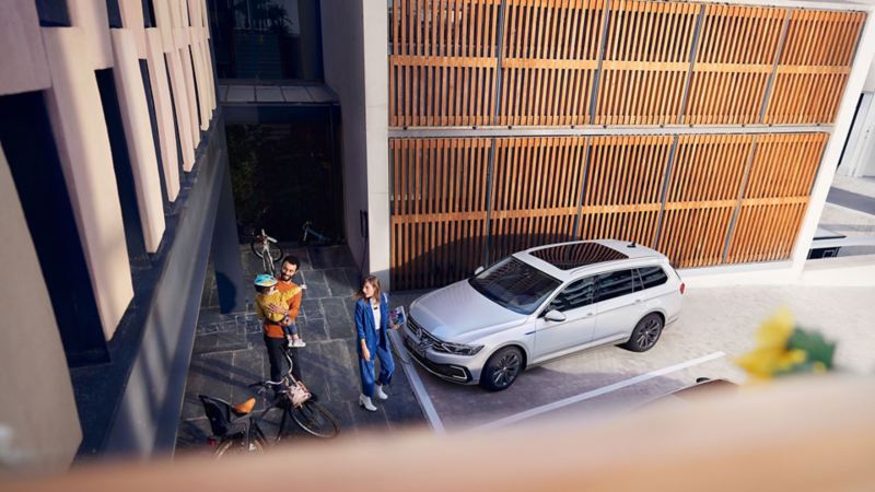 Vue extérieure d’en haut en diagonale de la VW Passat GTE Variant sur une place de stationnement, devant une paroi en bois, famille sur un trottoir