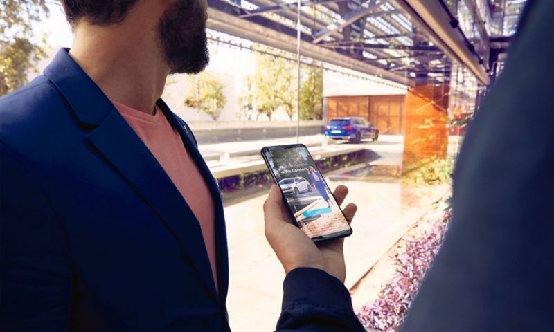 En person holder en mobiltelefon med WeConnect-appen i hånden, i baggrunden holder en VW Passat Variant parkeret.