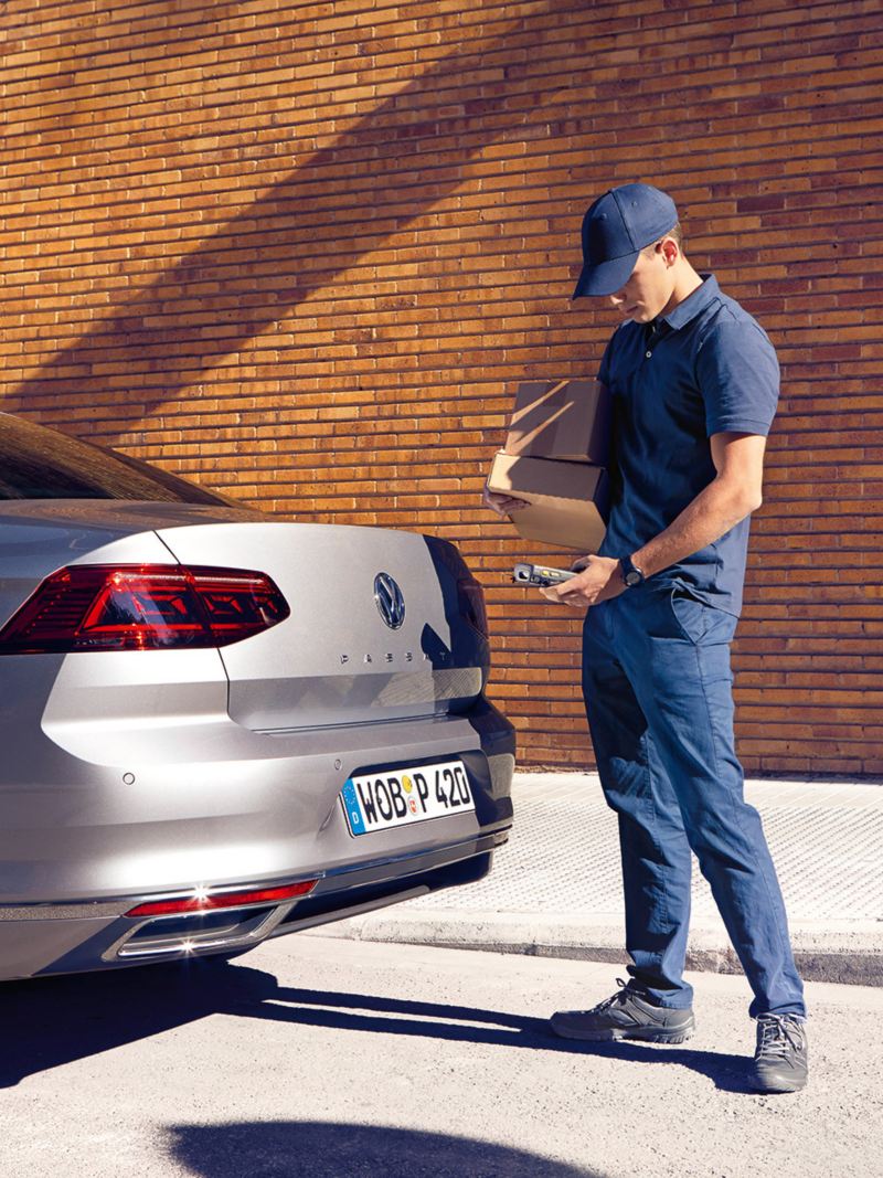 We Deliver: Deliverer stands at the rear of a Passat saloon and is about to open the luggage compartment flap with a smartphone.