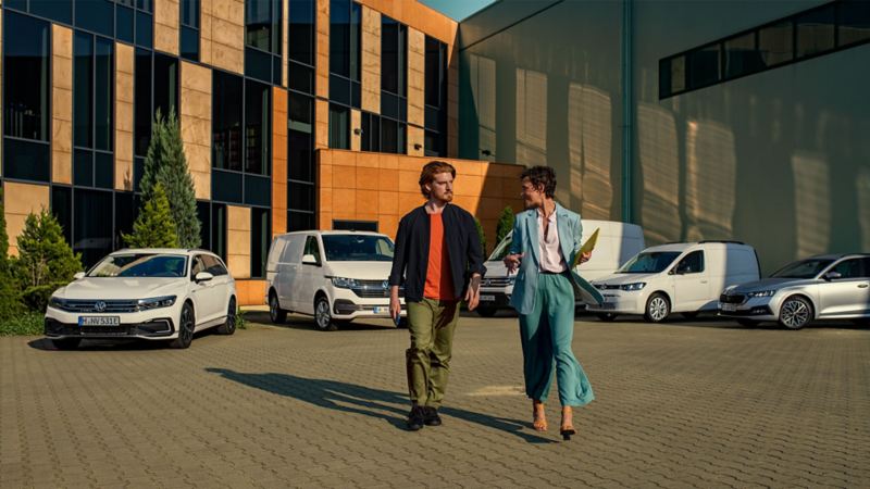 Frau und Mann gehen auf Kamera zu, im Hintergrund diverse Fahrzeuge des Volkswagen Konzerns vor Bürogebäude
