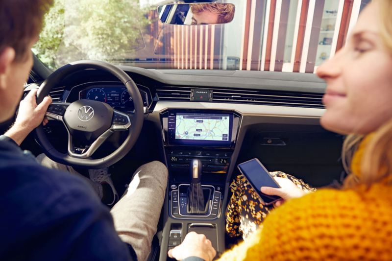 Coup d’œil par-dessus l’épaule d’un jeune homme et d’une jeune femme dans le cockpit de la Passat, au centre le système de navigation «Discover Pro», la femme tient un smartphone dans sa main.