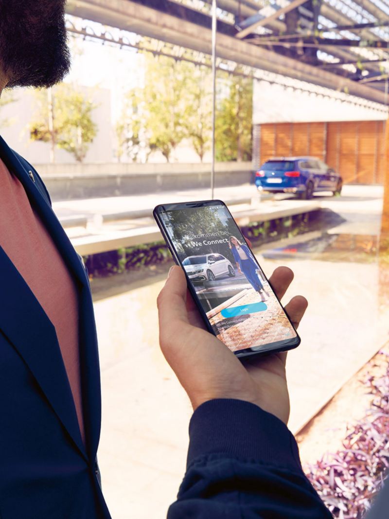 Un uomo tiene in mano il proprio smartphone con la schermata dell'app We Connect di Volkswagen. Fuori da una vetrata una Passat Variant in versione 'R-Line'.