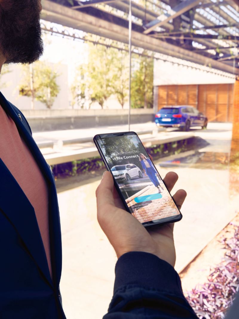 Une personne tient son smartphone avec l'application We Connect ouverte, une VW Passat SW en arrière-plan.