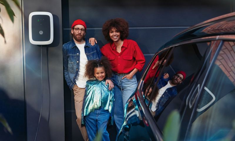 En mand, en kvinde og et barn står ved siden af en ID. Charger-wallbox, og i forgrunden en afskåret visning af en VW Passat GTE Variant.