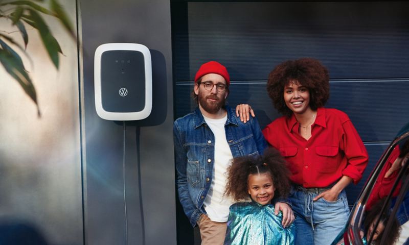 ID. Charger Wallbox an Wand neben Garage befestigt, Familie steht daneben und lächelt