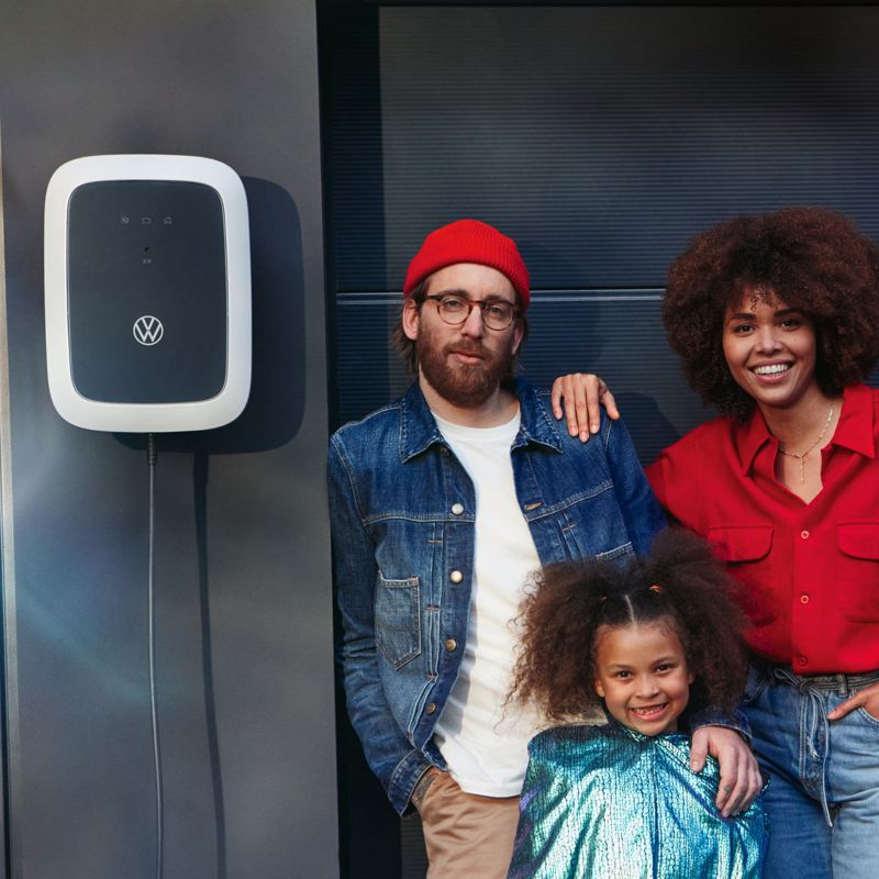 ID. Charger Wallbox an Wand neben Garage befestigt, Familie steht daneben und lächelt