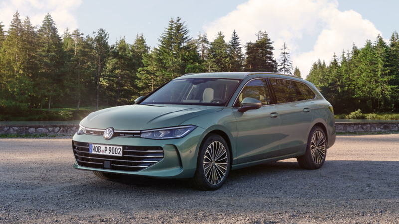 VW Passat vert bleutée stationnée sur une surface de gravier, la zone borde une forêt de pins.