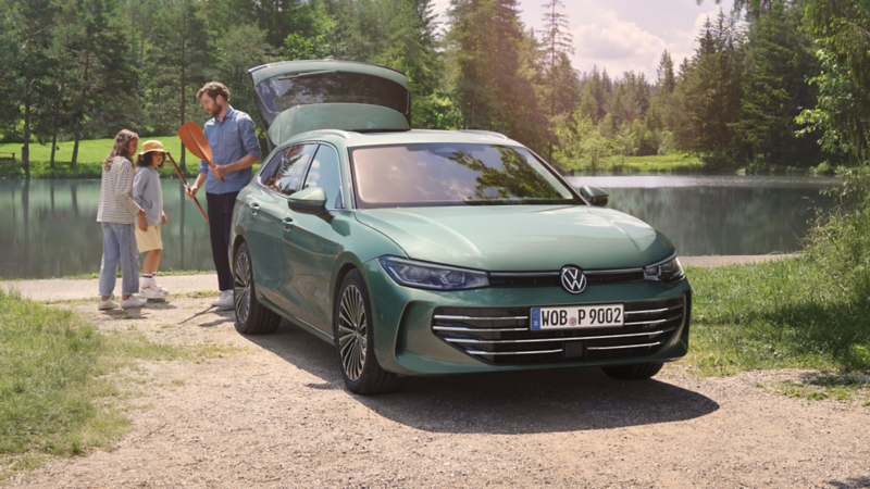 Zielononiebieski Volkswagen Passat z otwartym bagażnikiem stoi nad jeziorem. Mężczyzna rozdaje dwójce dzieci zabawki.