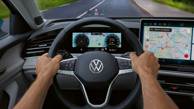 Vue de l’intérieur d’une VW Passat avec zoom sur le côté conducteur avec un grand écran couleur et l’affichage tête haute. Deux mains tiennent le volant.