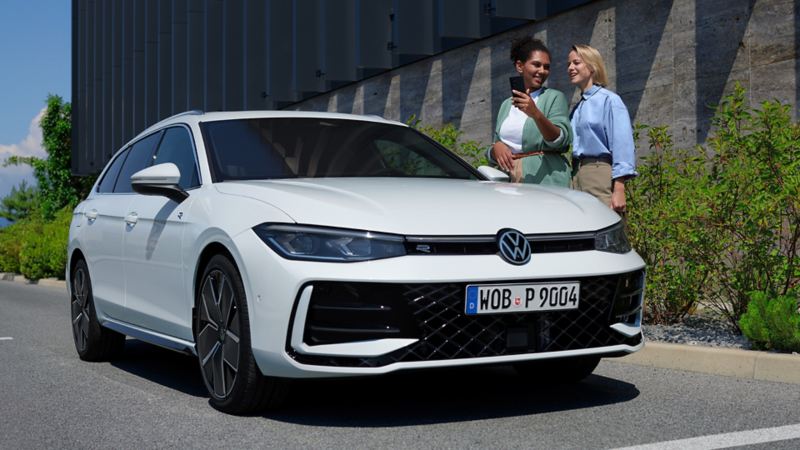 Een witte VW Passat parkeert dwars voor een gebouw, twee vrouwen staan aan de bestuurderskant erachter, één vrouw houdt haar smartphone vast.