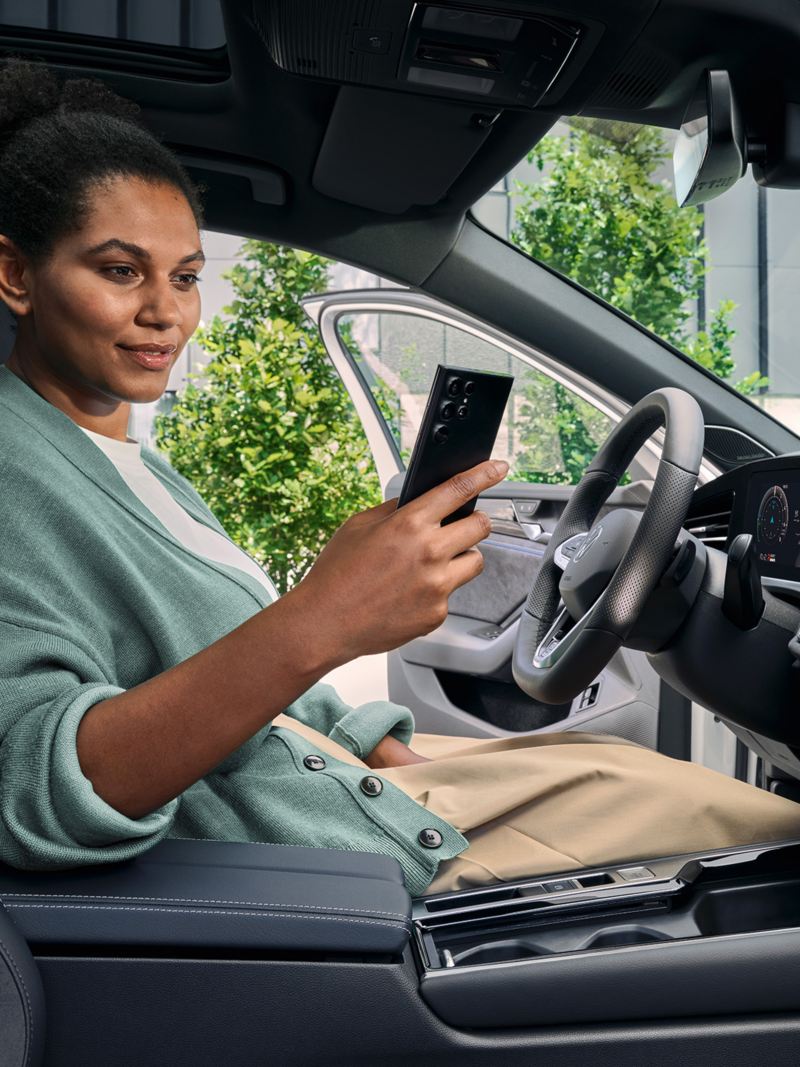 Een vrouw zit op de bestuurdersstoel van de geparkeerde VW Passat met de deur open en kijkt op haar smartphone.