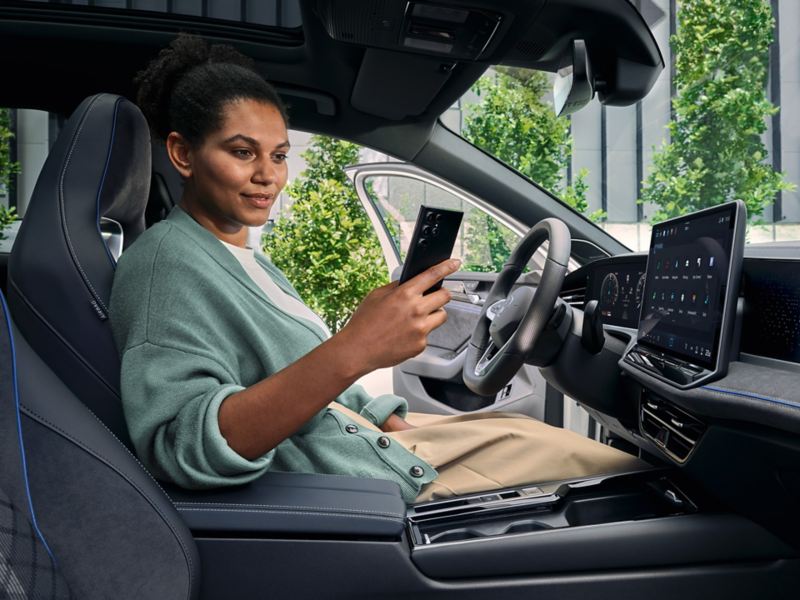 A woman sits in the driver seat of the parked VW Passat with the door open looking at her smartphone.