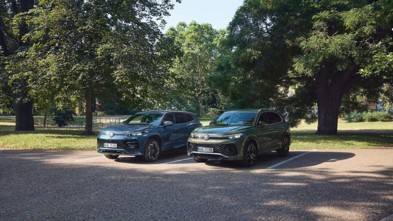 Ein blauer VW Tayron und ein grüner VW Tiguan stehen auf dem Parkplatz vor einer Parkanlage.