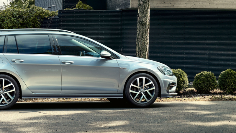 Silberner Volkswagen Golf vor schwarzer Backsteinmauer geparkt, neben einem Baum und umgeben von grünen Büschen.