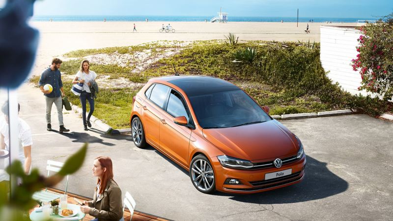 Una coppia di ragazzi si avvicina ad una Volkswagen Polo, vista 3/4 frontalmente, parcheggiata a bordo spiaggia.