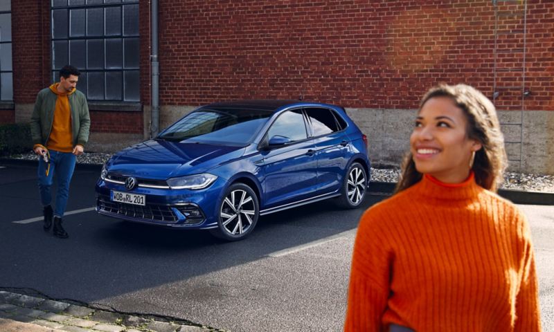 Une Volkswagen Polo bleue est garée dans un parking, un homme se tient devant elle et regarde le capot.