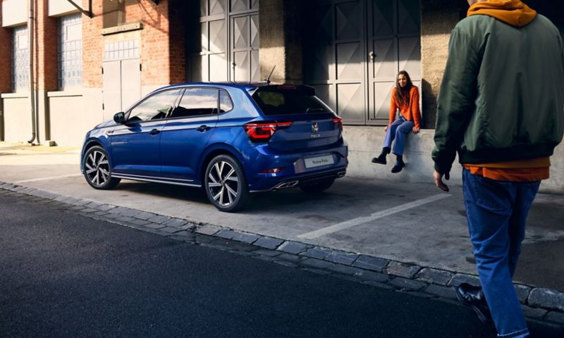 Un ragazzo si avvicina a Volkswagen Nuova Polo, vista 3/4 posteriormente, parcheggiata al lato di una strada con una ragazza seduta accanto.