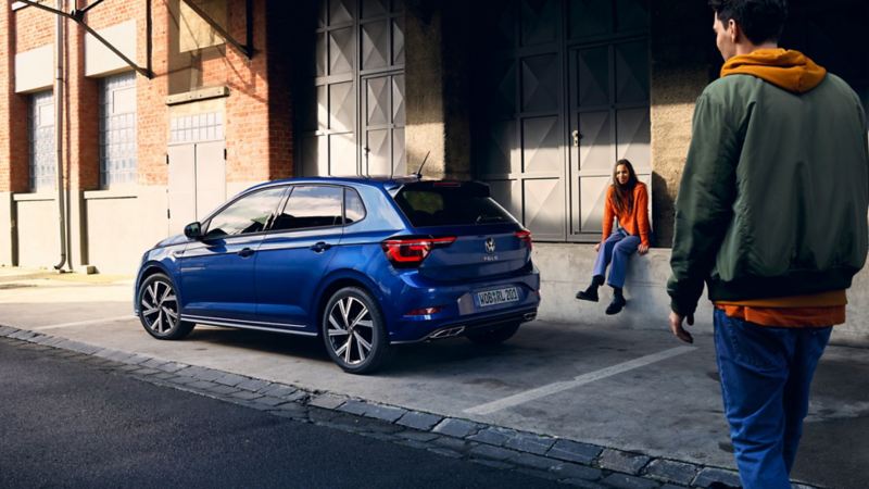 Une VW Polo bleue visible devant une rampe, un homme passe devant l’arrière doté des feux arrière à LED en option.