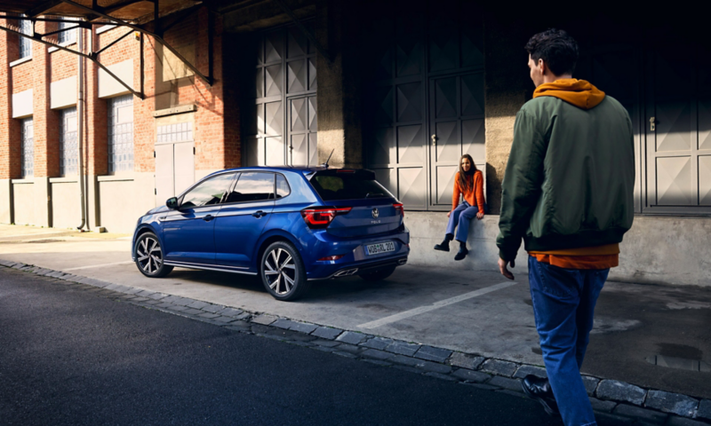 Un ragazzo si avvicina a Volkswagen Nuova Polo, vista 3/4 posteriormente, parcheggiata al lato di una strada con una ragazza seduta accanto.