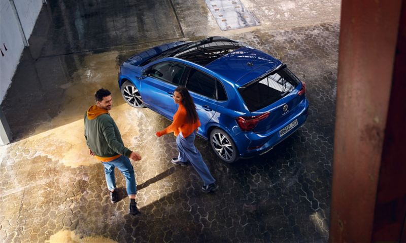A blue VW Polo with an optional panoramic sliding roof is parked in a hall, a couple is walking towards it.