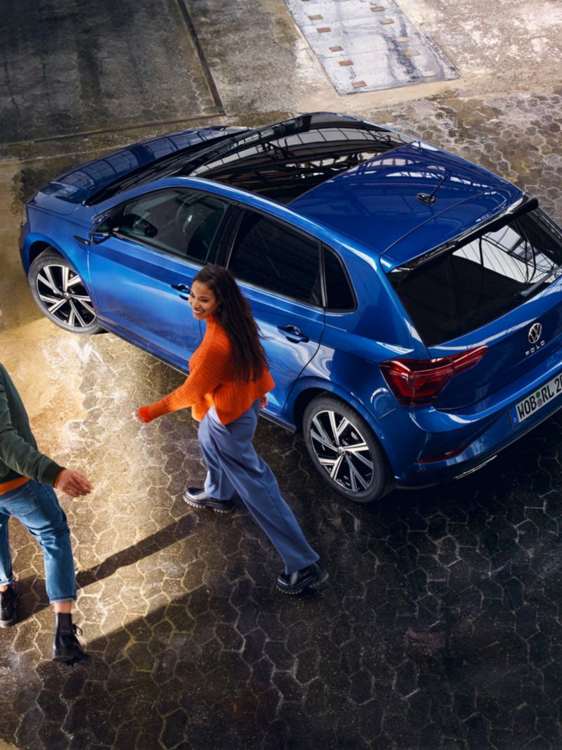 Un couple se dirige vers une Polo Volkswagen bleue avec toit coulissant panoramique à l’arrêt dans une halle.