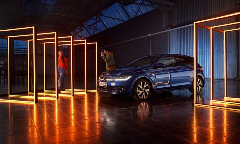 Une Volswagen Polo bleue avec signature lumineuse LED et phares matriciels LED se tient dans un hall sombre entouré d'éléments lumineux rectangulaires.
