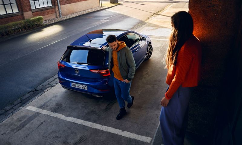A new blue Polo with tinted rear windows is parked in front of a ramp, a woman is sitting on the ramp, a man is walking towards it. 