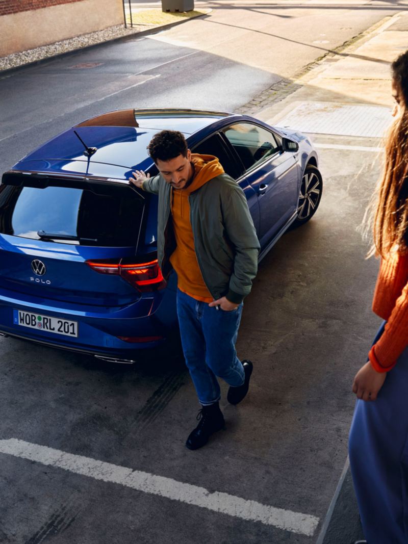 A new blue Polo with tinted rear windows is parked in front of a ramp, a woman is sitting on the ramp, a man is walking towards it. 