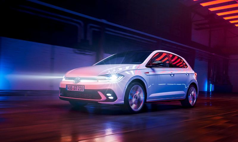 Une VW Polo GTI blanche roule dans une halle avec des phares matriciels à LED allumés et une barre lumineuse à l’avant.