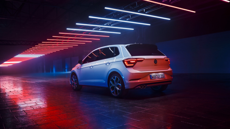 Blick von hinten auf einen weißen Polo GTI, welcher mit eingeschalteten LED-Heckleuchten in einer dunklen Halle unter Leuchtelementen steht.
