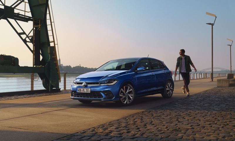 Ein blauer VW Polo steht auf einem Hafengelände, Blick schräg auf die Front. Ein Mann geht am Heck vorbei. 