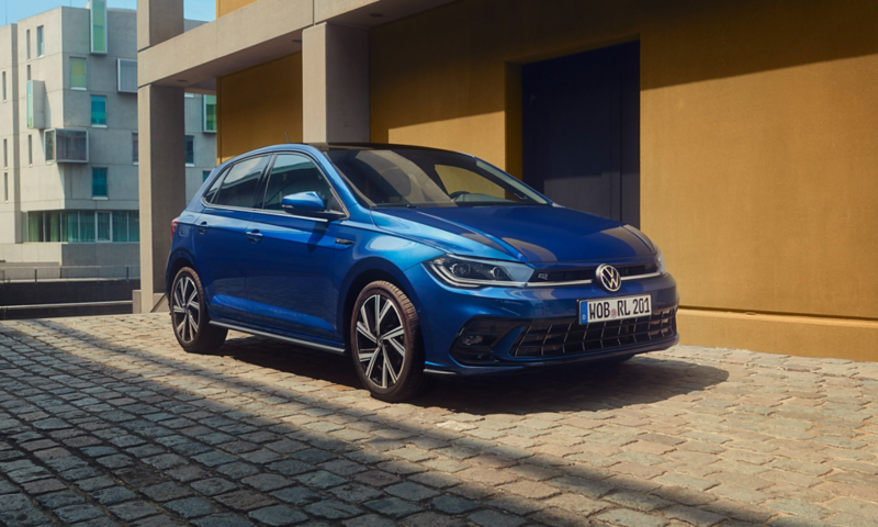 A blue Polo R-Line parked on cobblestones next to a building. View of the front and passenger side. 