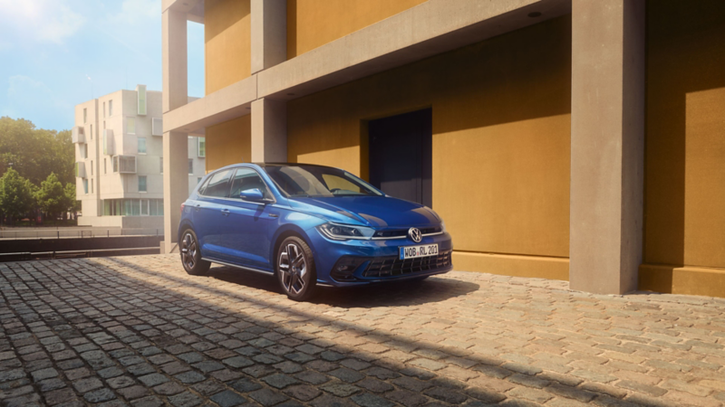 Ein blauer Polo R-Line parkt auf Kopfsteinplaster neben einem Gebäude. Ansicht der Front und Beifahrerseite. 