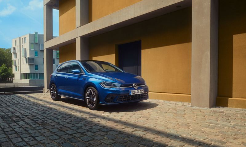 A blue Polo R-Line is parked on cobblestones next to a building. Front and passenger side view.