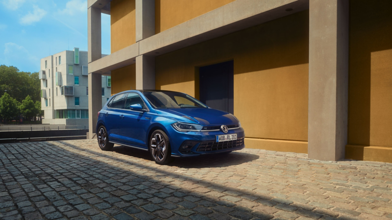 Ein blauer Polo R-Line parkt auf Kopfsteinplaster neben einem Gebäude. Ansicht der Front und Beifahrerseite. 