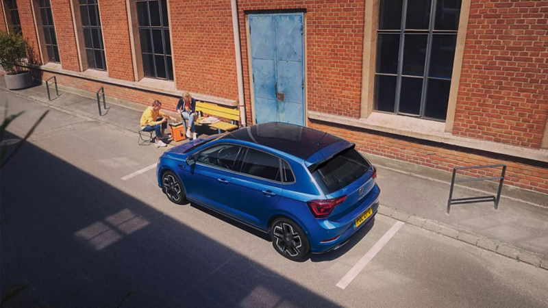 View from diagonally above of a parked blue Polo R-Line with roof pack. A couple is playing chess next to it.